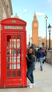 JoJo from JoJo's Cup of Mocha, a solo black female traveler standing in front of the iconic red phone booth, in front of Big Ben, in London.